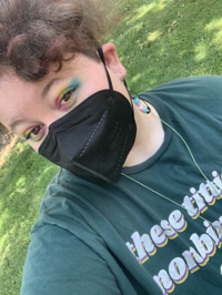 Selfie of Ci, a white fat person, is wearing a black mask with pan flag colored eyeshadow, a shirt that says “these titties nombinary too”. He also has on trans safety-pin earrings. 